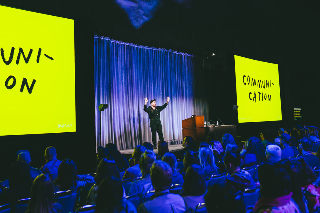 Photo of Adam speaking to a crowd. Slideshow displayed beside him is one handwritten word – COMMUNICATION – on yellow background.