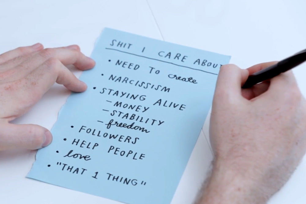 Image from online course. Adam's hands writing a list on blue paper. Page reads as follows. Headline: Shit I Care About followed by a list of bullet points. Need to create. Narcissism. Staying alive, sub list is money, stability, freedom. List continues.