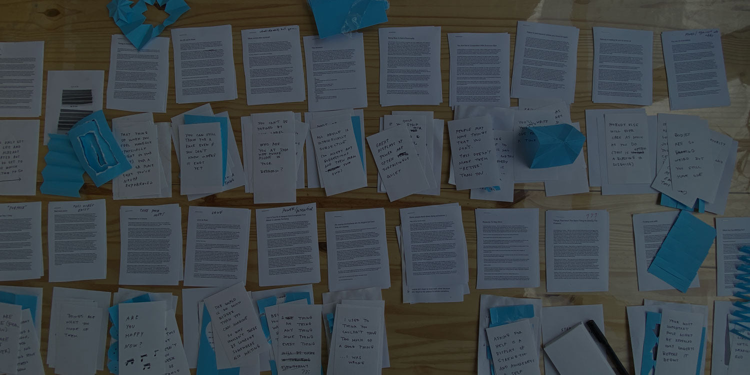 A photo of Adam's desk covered in printouts of text too small to read with handwritten notes and blue folded paper artwork strewn about.